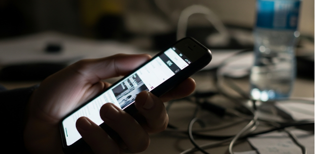 A hand holds a smartphone displaying articles about PR for startups and small businesses, with cables and a water bottle nearby.